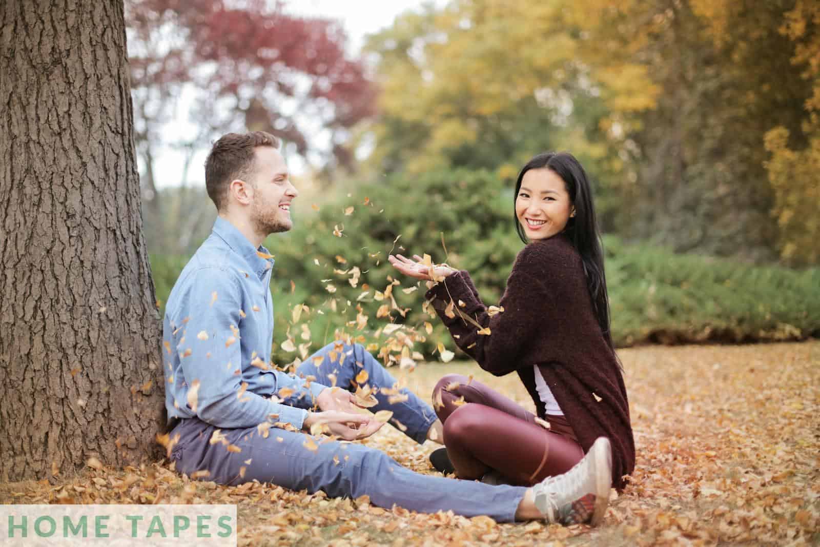 Side view of cheerful couple in casual clothes sitting in park and having fun while scattering dry leaves on autumn day